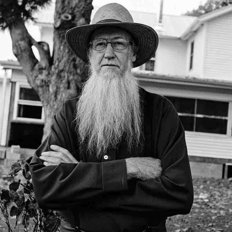 The black and white image shows an Amishr man with a long, white beard, wearing a hat and glasses. He has his arms crossed and is looking directly at the camera. He appears to be standing in front of a building with a tree to his left. The overall tone of the image is serious and Amish tradition.